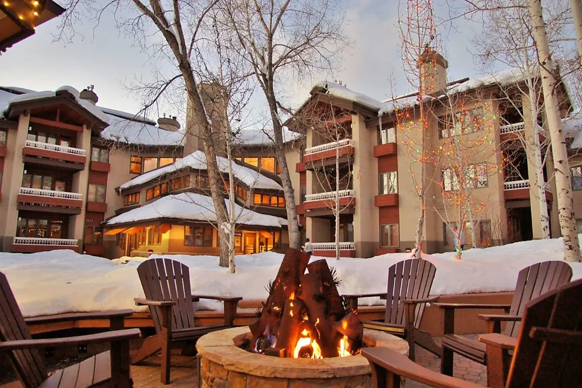 A warmly lit exterior scene featuring a fire pit surrounded by inviting chairs, set against a backdrop of snow-covered ground, twinkling lights on winter trees, and the exterior of the resort building. Small groups gather, enjoying hot beverages and lively conversation while basking in the glow of the fire.