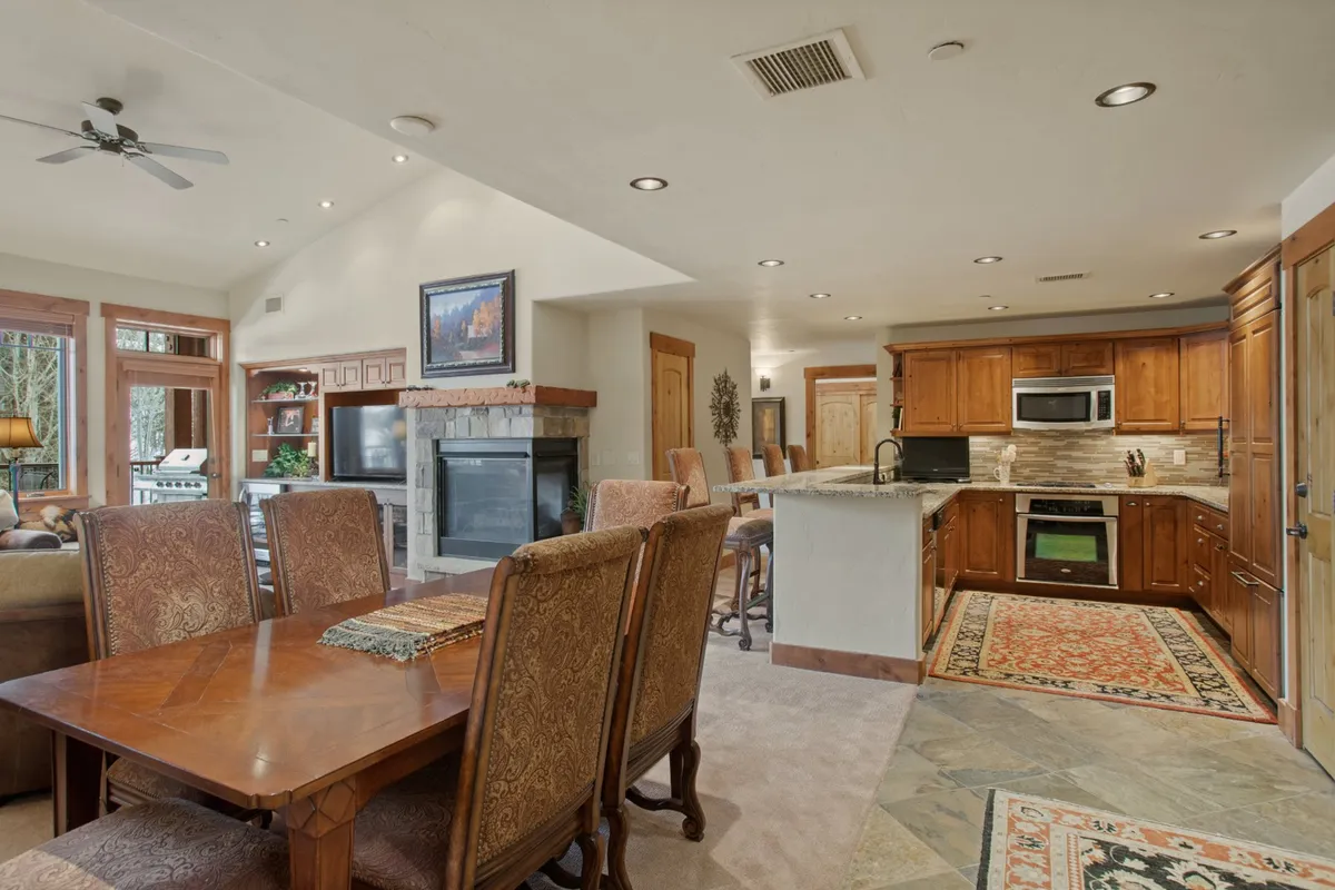 An inviting open-concept area combining the dining table, fireplace, kitchen, and a cozy seating area. The layout is designed for social interaction, family meals, and entertaining guests, all set within a sunlit, vaulted space.