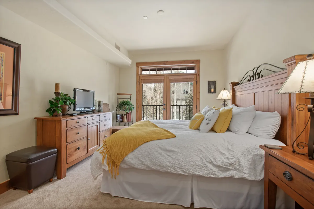 A well-appointed bedroom featuring a plush bed with cheerful yellow accents, wooden furniture, a smart TV, and doors leading to a balcony. This space blends comfort with a touch of elegance and natural light.