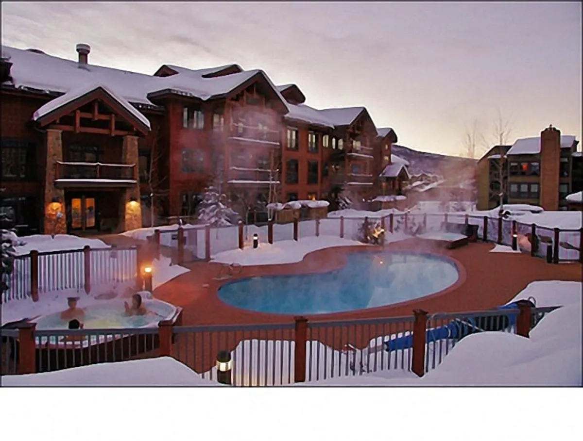 A tranquil winter exterior with multiple outdoor hot tubs and a swimming pool, all enveloped by snow and set against the backdrop of the resort’s mountain architecture. Guests are seen relaxing in the steamy hot tubs, enjoying the serene winter landscape.
