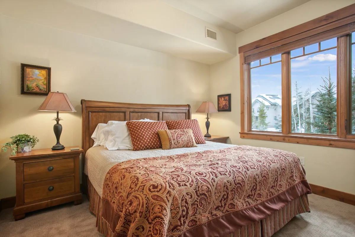 A spacious bedroom with an ornate bed dressed in luxurious bedding, a window offering mountain views, and a cozy nightstand with a lamp. The natural light creates a tranquil, welcoming atmosphere ideal for relaxation.