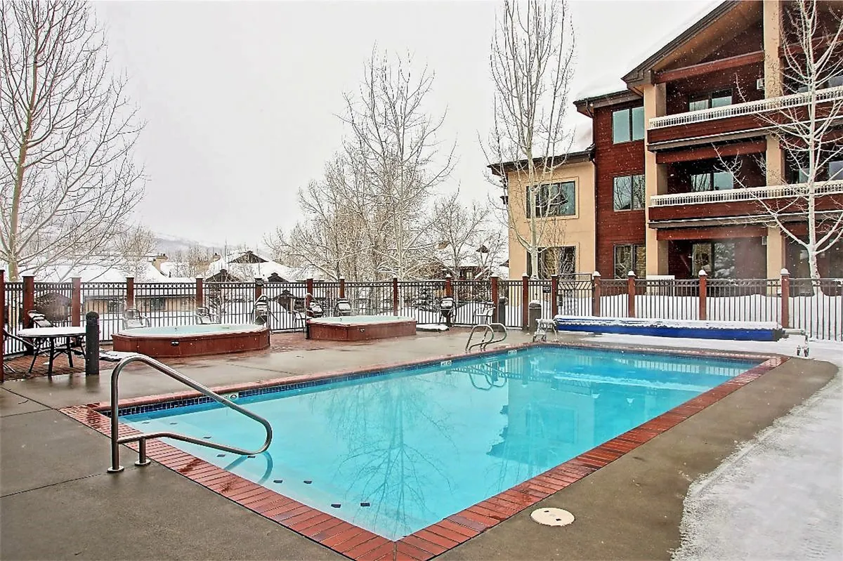 This image displays a serene exterior scene of a swimming pool and hot tub surrounded by trees and snow, with a view of the building facade. The steaming hot tub contrasts with the wintry backdrop, illustrating year-round outdoor amenities.