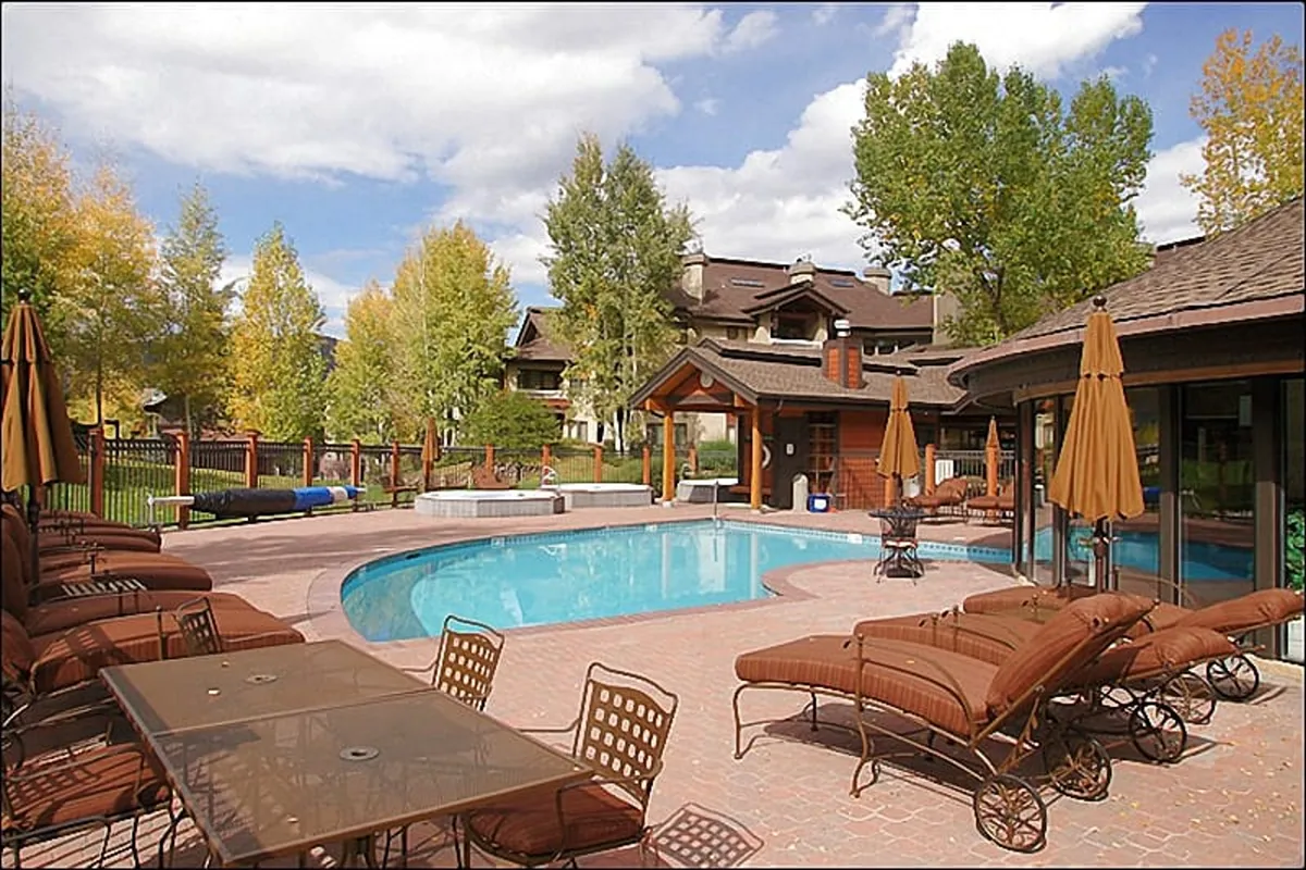 A vibrant outdoor pool area with lounge chairs, a dining table, and surrounding greenery, set against the backdrop of the lodge buildings. The space is inviting for families to gather, swim, and relax during warmer months.