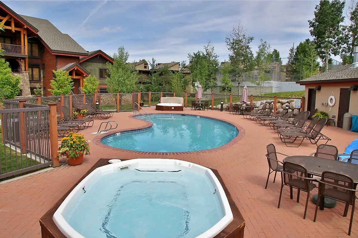An exterior view of a sparkling pool and hot tub, bordered by lounge chairs and a dining area, set amid manicured landscaping. The inviting scene suggests both daytime sunbathing and evening relaxation by the water.
