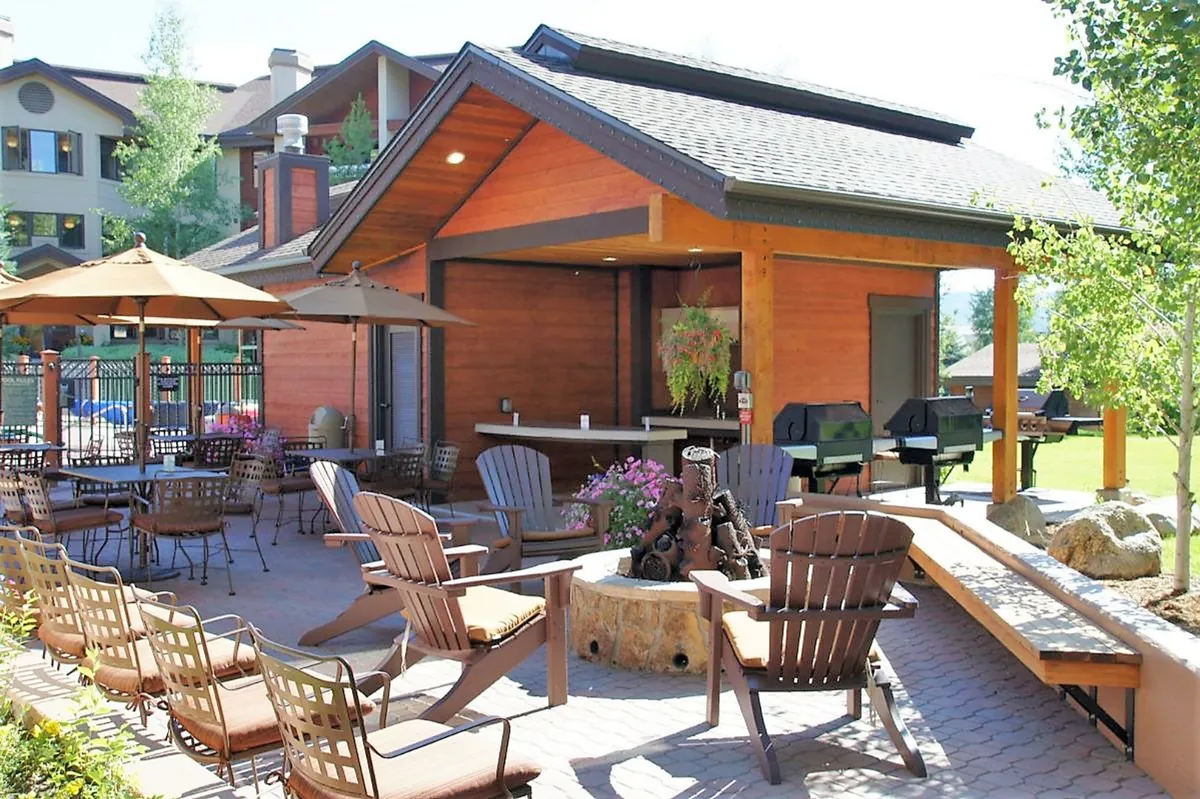 A cozy outdoor seating area with a fire pit, grill area, bar counter, and a landscaped garden. The space is designed for social gatherings, relaxation, and outdoor dining, set against a backdrop of lush greenery.