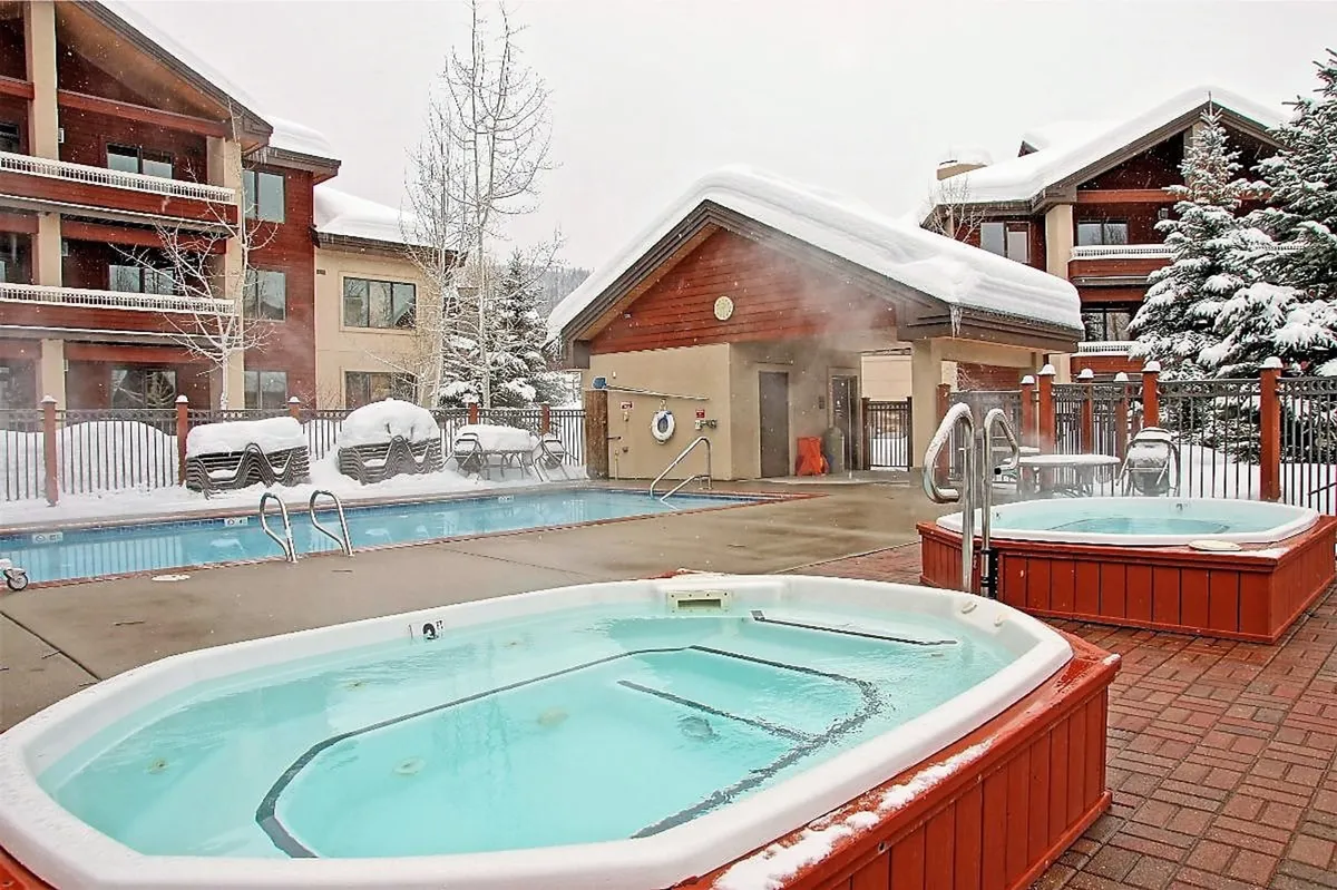A wide exterior view of outdoor hot tubs and a swimming pool nestled among snow-covered trees, with the resort building in the background. Guests soak in the hot tubs as dusk settles, exchanging stories and taking in the peaceful, wintry setting.