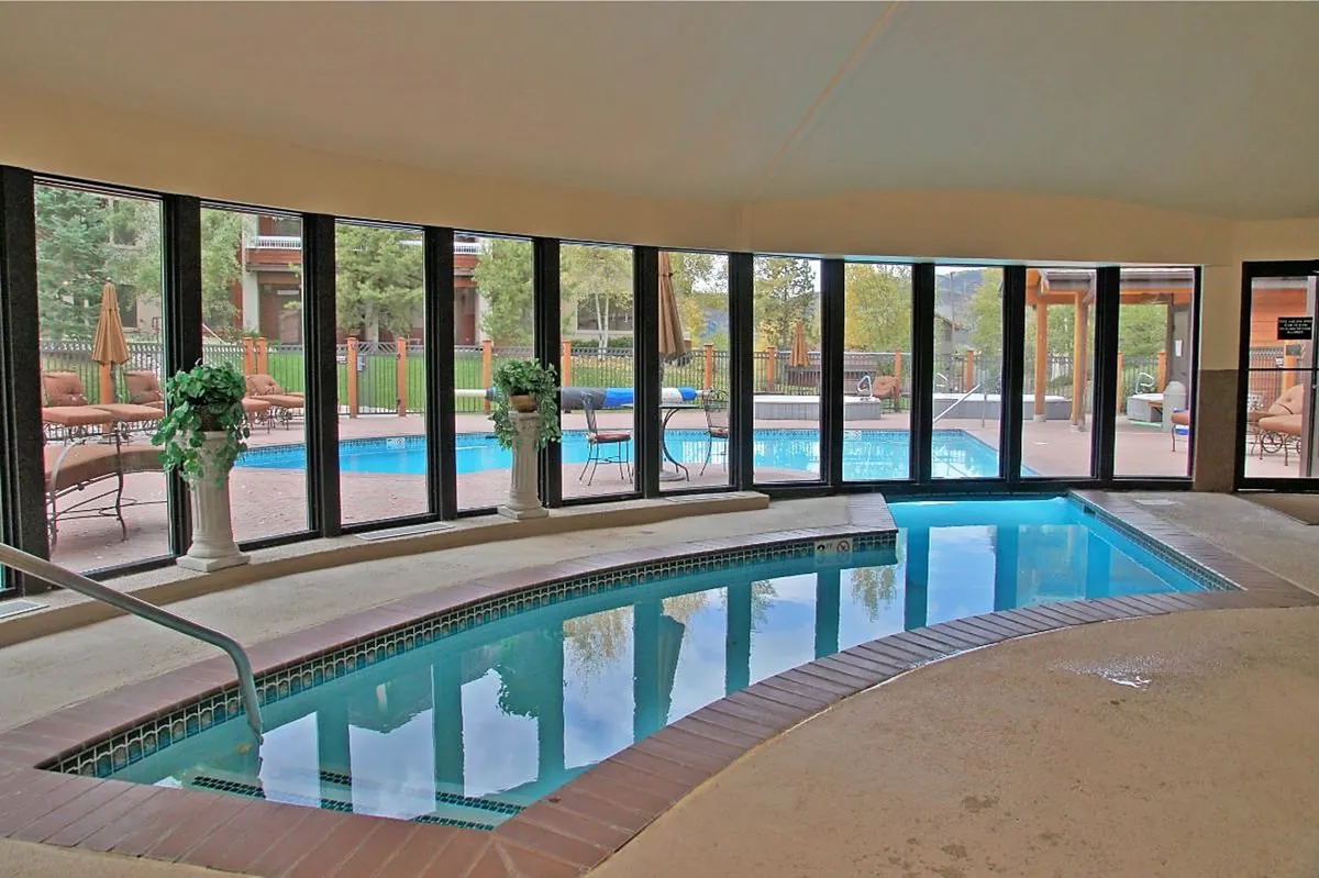 A tranquil indoor hot tub area with expansive windows looking out to the outdoor pool. Decorative plants soften the space, and the inviting water reflects the natural light, creating a serene retreat for relaxation and socializing.