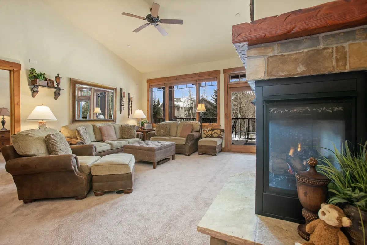 A warm and welcoming living room featuring a comfortable sectional sofa, a crackling fireplace, and large windows that provide expansive ski resort and mountain views. The room is bathed in natural light, with plush furnishings arranged for relaxation and conversation.